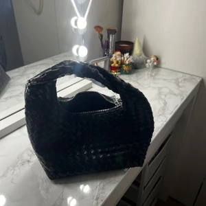 Svart flätad handväska - Snygg svart handväska i flätat mönster med dragkedja. Perfekt för både vardag och fest! Den har en unik design med ett bekvämt handtag och är tillverkad i ett stilrent material som ger en lyxig känsla.