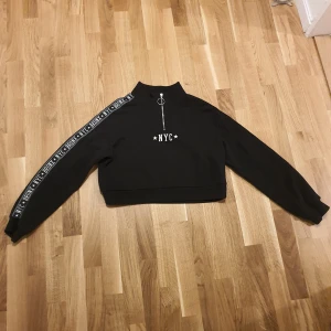 Svart croppad tröja från H&M Divided - Säljer en svart croppad tröja från H&M Divided med texten 'NYC' på bröstet. Tröjan har en dragkedja med ringdetalj och text längs ärmarna. Perfekt för en avslappnad stil.