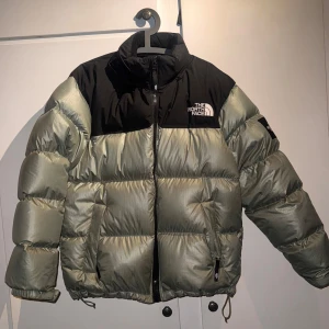 Grön och svart dunjacka från The North Face - Säljer en grön och svart pufferjacka från The North Face. Jackan har en hög krage och dragkedja framtill. Den är perfekt för kalla dagar och har märkets logga på bröstet och ärmen. Strl XL. Går att dra år nedtill med dragsko. Har en diskret fläck på ena armen. 