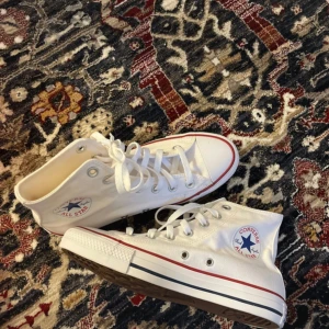 Vita Chuck Taylor converse - Säljer ett par klassiska vita Converse All Star sneakers med hög snörning och röd/blå randdetalj på sulan. Perfekta för en avslappnad stil. Aldrig använda endast testade💖