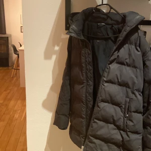 Svart pufferjacka med dragkedja - Säljer en svart pufferjacka med dragkedja och hög krage. Jackan har en quiltad design och en logotyp på ärmen. Perfekt för kyliga dagar. Pris kan diskuteras 