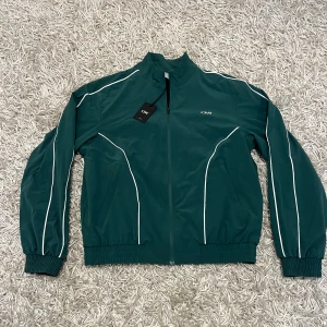 ICIW träningströja - Säljer en snygg grön vindjacka från ICIW i nytt skick. Jackan har en dragkedja framtill och vita detaljer längs ärmarna. Perfekt för vår och höst! Den är långärmad och har en normal passform. Perfekt för träning eller en casual look.