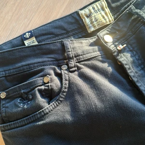 Svarta jeans från Morris - Säljer ett par snygga mörklbåa byxor från Morris i storlek 32/32. De har en klassisk femficksdesign med knappar och en läderpatch med logga baktill. Perfekta för både vardag och fest! Jag är 179cm lång och väger 77-78kg. Byxorna är tvättade på kemtvätt inför försäljningen