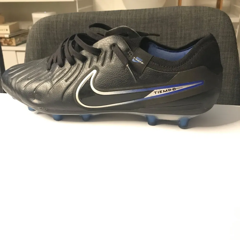 Säljer ett par svarta Nike Tiempo fotbollsskor i mycket bra skick. Skorna har en snygg design med vita och blå detaljer samt snörning för perfekt passform. Perfekta för fotbollsspel på gräsplaner. Sulan har blå dobbar för bra grepp.. Skor.