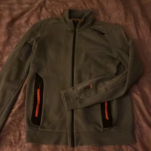 Grå fleecetröja från Sail Racing - Säljer en grå fleecetröja från Sail Racing i storlek S. Tröjan har en dragkedja framtill och två fickor med orange detaljer. Perfekt för kyliga dagar och har en sportig look. Passar bra till både vardag och utomhusaktiviteter.         PRISET GÅR ATT DISKUTERA 🤍🩶🤍🩶