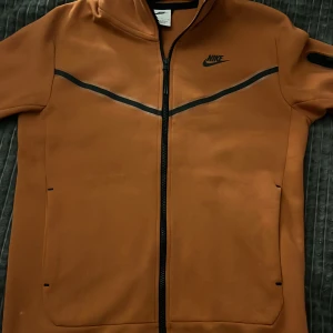  Nike Tech Fleece i orange färg - Säljer en snygg brun Nike Tech Fleece i storlek S. Den har en dragkedja framtill och är perfekt för höst och vår. Jackan är i okej skick. Lite fläckig på armen, samt där etiken ligger och vid zippern. Mer orange i färgen än brun det är bara ljuset. Pris runt 1499 kr köpt för 1099 kr mitt pris = 349 kr.
