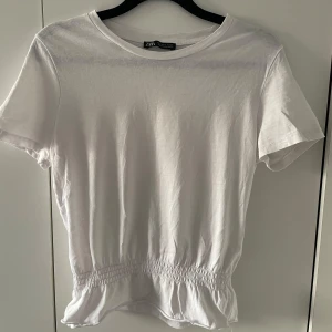 Vit topp från Zara - Säljer en söt vit topp från Zara i bomull. Den har korta ärmar och en smockdetalj i midjan som ger en fin volangkant. Perfekt för sommardagar eller att styla med en jeansjacka för en avslappnad look. 🕊️