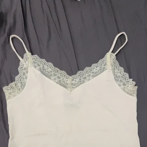 Vit linne med spetsdetaljer - Säljer ett superfint vitt linne med tunna axelband och vacker spetsdetalj vid halsringningen. Linnet är i bra skick och har en mjuk och bekväm passform. (smutsvit och ej tandbleksvit färg)