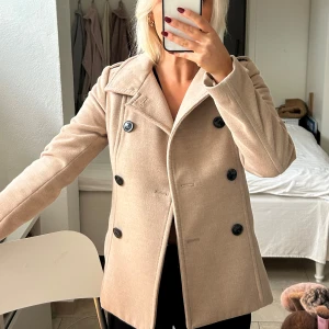 Beige kappa med knappar Stl 34 - Elegant beige kappa med dubbelknäppning och klassisk krage. Perfekt för höst och vår, med en stilren design som passar till många tillfällen.