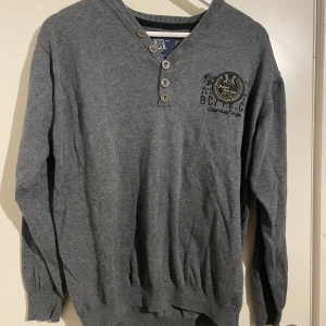 Grå tröja från Napapijri - Säljer en stilren grå tröja från Napapijri med knappar vid halsen och ett tryck på bröstet. Perfekt för en avslappnad look. Tröjan har långa ärmar och ribbade muddar.