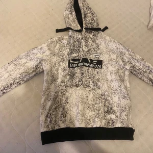  Emporio Armani Hoodie - Snygg vit och svart hoodie från Emporio Armani med ett unikt mönster. Den har en stor ficka framtill och justerbar huva med svarta snören. Perfekt för en avslappnad stil.