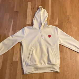 Vit hoodie från Comme des Garçons - Säljer en stilren vit hoodie från Comme des Garçons med det ikoniska röda hjärtat broderat på bröstet. Tröjan har en klassisk passform med långa ärmar och en bekväm huva. Perfekt för en avslappnad look.