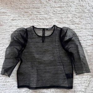 Svart transparent blus från Gina Tricot - Snygg svart transparent blus från Gina Tricot med puffiga trekvartsärmar. Perfekt för en stilren och trendig look. Blusen har en rund halsringning och knäppning i nacken.