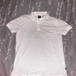 Hugo Boss pikétröja  - Säljer en klassisk vit pikétröja från BOSS i storlek S. Tröjan är i nytt skick med etikett kvar och har en stilren design med krage och knappar framtill. Perfekt för både vardag och festliga tillfällen. Materialet är mjukt och bekvämt, vilket gör den idealisk för sommarsäsongen.
