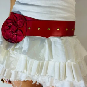 Rött bälte med blommig detalj - Snyggt rött bälte med en stor blommig detalj på sidan och guldfärgade nitar. Perfekt för att ge en färgklick till din outfit. Bältet är brett och ger en fin siluett. 💞105 cm