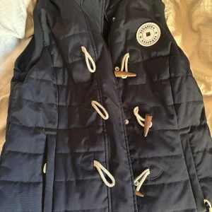 Svart quiltad väst från Redgreen - Säljer en snygg svart quiltad väst från Redgreen med dekorativa knappar och en rund logotyp på bröstet. Perfekt för kyligare dagar och ger en stilren look. Västen har en klassisk design med praktiska detaljer. Har aldrig vart använd då min pappa köpte den till mamma men hon har ej hunnit använda. Prislappen är kvar, perfekt julklapp🤗