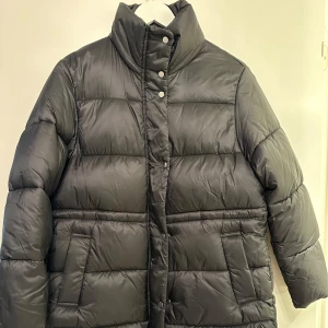 Mango Svart dunjacka - Säljer en svart mango dunjacka med hög krage och knäppning framtill. Jackan har en quiltad design och långa ärmar, perfekt för kalla dagar. Den har både dragkedja och knappar för extra skydd mot kyla. Str m men passar en s också använd ett par gånger men i fin skick 