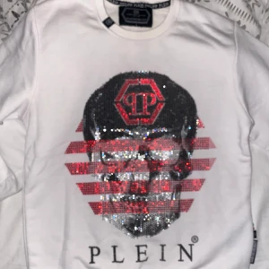 Vit tröja från Philipp Plein - Säljer en cool vit tröja från Philipp Plein med ett stort, glittrigt dödskallemotiv och röda detaljer på framsidan. Baksidan har också ett dödskalletryck med texten 'PHILIPP PLEIN 1978'. Perfekt för att sticka ut på festen eller bara ha en chill dag. Tröjan är i bra skick och redo för nya äventyr! L i junior storlek så passar er med s skulle ja säga.