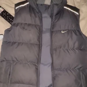 Svart dunväst från Nike - Säljer en svart dunväst från Nike i bra skick. Västen har en dragkedja framtill och reflekterande detaljer på axlarna. Perfekt för höst och vår när du vill hålla dig varm men ändå se sportig ut. Passar bra till både träning och vardagsbruk. Pris kan diskuteras