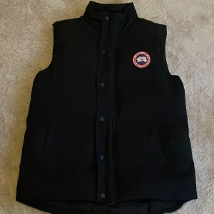 Svart dunväst från Canada Goose - Säljer en svart dunväst från Canada Goose i mycket bra skick. Västen har en dragkedja och knappar framtill samt en broderad logotyp på bröstet. En stor innerficka samt en innerficka med dragkedja.  Perfekt för höst och vinter, håller dig varm och stilren. Passar bra till vardagligt användande. Storlek M passar även S