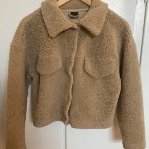 Mysig beige teddyjacka i mycket bra skick. Perfekt för höst och vinter med sin varma och gosiga stil. Jackan har långa ärmar och en klassisk krage. Den är croppad och har en avslappnad passform. Perfekt för att hålla sig varm och trendig!