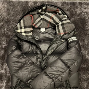 Burberry jacka  - Snygg svart dunjacka från Burberry med klassiskt rutigt foder i huvan. Jackan har dragkedja och knappar framtill samt två fickor med dragkedja. Perfekt för kyliga dagar med stil.