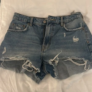 Blå jeansshorts från Zara - Säljer ett par coola blå jeansshorts från Zara med slitna detaljer och fransiga kanter. Perfekta för en avslappnad sommarlook. De har en klassisk femficksdesign och knappgylf.