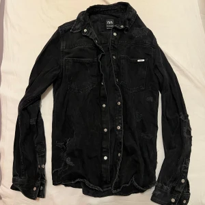 Svart jeansskjorta från Zara - Säljer en svart jeansskjorta från Zara i avslappnad stil. Skjortan har slitna detaljer och knappar framtill. Perfekt för en cool och avslappnad look. Passar bra till både vardag säljs för 80