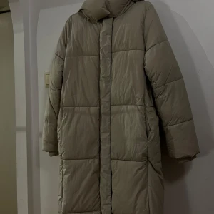 Beige dunjacka från Pull&Bear - Säljer en supermysig beige dunjacka från Pull&Bear i storlek M. Perfekt för kalla vinterdagar med sin långa design och varma vaddering. Jackan har en dragkedja framtill och långa ärmar. Den är i mycket bra skick och redo för nya äventyr!