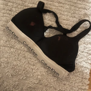 Svart sport-bh från Calvin Klein - Säljer en svart sport-bh från Calvin Klein. Den har en bred resår med loggan i vitt och svart. Perfekt för träning eller vardagsbruk. Den är i bra skick och superbekväm! Har råkat få lite blekning på den därav missfärgningen