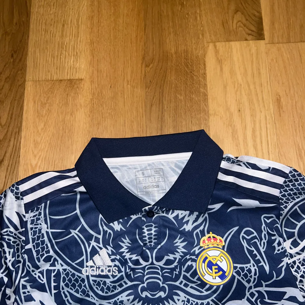 Säljer en supercool blå Real Madrid fotbollströja från Adidas med ett unikt drakmönster i vitt. Tröjan har korta ärmar och en klassisk krage. Perfekt för alla fans som vill sticka ut på läktaren eller planen! 🐉⚽köpte den för en vecka seda, har bara haft på mig den en gång, gick inte att returnera, behöver pengarna tillbaka därför säljer jag den, pris går att diskutera vid snabb affär. T-shirts.