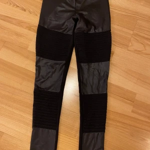 Svarta leggings från Rainbow - Snygga svarta leggings från Rainbow i storlek 36/38. De har en cool ribbad detalj på knäna och är gjorda i ett glansigt syntetmaterial som ger en läderliknande look. Perfekta för en kväll ute eller en avslappnad dag med stil. 🖤