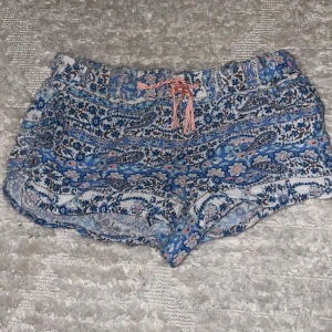 Blommiga shorts med knytband - Säljer ett par supersköna blommiga shorts i blått och rosa med ett snyggt knytband i midjan. Perfekta för sommardagar på stranden eller bara chilla hemma. De är i bra skick och har en lös passform som gör dem extra bekväma. 🌸