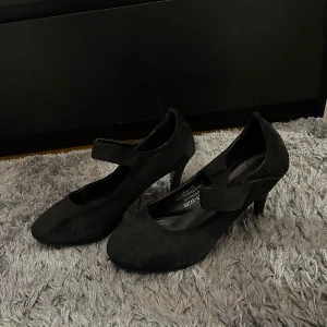 Svarta pumps från Lesara - Säljer ett par eleganta svarta pumps från Lesara i storlek 41. Skorna är i mocka och har en bekväm klack. Perfekta för festliga tillfällen eller en utekväll. De har en stilren design med en rem över foten för extra stöd.