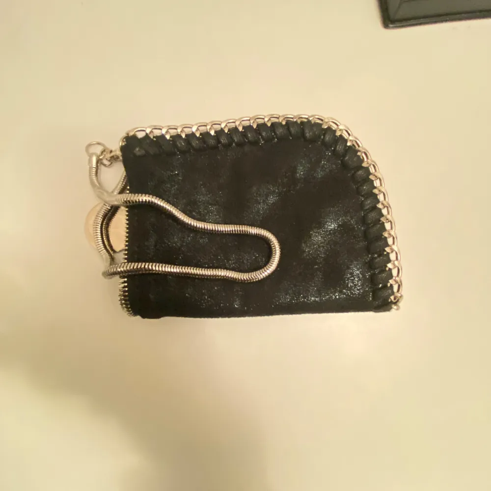 Säljer en snygg svart plånbok med coola kedjedetaljer runt kanterna. Den har en unik design och är perfekt för att hålla dina kort och kontanter säkra. Passar perfekt i väskan eller fickan. Perfekt accessoar för både vardag och fest!. Laukut & Käsilaukut.