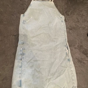Ljusblå jeansklänning - Säljer en snygg ljusblå jeansklänning med axelband och dragkedja bak. Perfekt för sommardagar eller en avslappnad vårlook. Klänningen har en enkel och stilren design som gör den lätt att matcha med olika accessoarer.