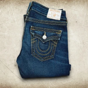 Mörkblå jeans från True Religion - Säljer ett par snygga mörkblå jeans från True Religion. De har en cool design med kontrasterande sömmar och en unik fickdetalj med knapp. Perfekta för en avslappnad men stilren look. Passar både till vardags och fest!