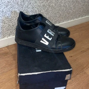 Versace skor  - Snygga svarta sneakers från Versus Versace i skinn med breda remmar över foten där det står 'VERSUS'. Perfekta för en stilren look, både till vardags och fest. Skorna är i mycket bra skick och kommer med originalkartong.