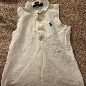 Vit ärmlös pikétopp från Ralph Lauren - Säljer en klassisk vit ärmlös pikétopp från Ralph Lauren. Den har en liten krage och knappar framtill, med den ikoniska loggan broderad i blått på bröstet. Superfin och stilren