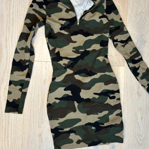 Camouflage klänning från H&M  - Säljer en cool camouflage klänning från H&M i storlek XS. Den har en dragkedja framtill och är långärmad, perfekt för en avslappnad stil. Materialet är mjukt och bekvämt, vilket gör den idealisk för höst och vår. Passar perfekt för en casual look eller en dag ute i naturen.