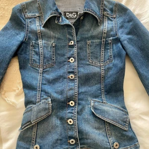 Jeansjacka från D&G - Säljer en snygg jeansjacka från D&G i klassisk blå denim. Jackan har en figursydd passform. Mått midja som bild 5, är  33 cm. Mått från armhåla till handled är  49 cm. Mått över byst  38cm. Står ingen storlek så XS är min gissning. 