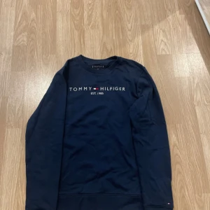 Tommy Hilfiger tröja - En klassisk tommy hilfiger tröja som är marin blå. Texten på överdelen är vit och tröjan har en liten tommy loggo även på högra arm vid handleden. Den är perfekt till hösten och skulle passa alla! Från storlek xs-m (Unisex)