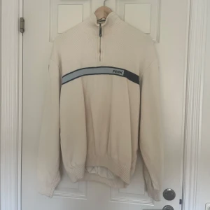 Beige tröja med dragkedja från Peak - Säljer en snygg beige tröja från Peak med en dragkedja vid halsen. Den har en mörkblå rand över bröstet med märkesnamnet. Perfekt för kyliga dagar!