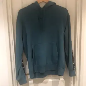 Säljer en snygg blå hoodie från Nike. Den har en stor ficka fram och en luva med dragsko. På ärmarna finns det coola detaljer och på ryggen står det 'NIKE' i svart. Perfekt för chill dagar eller träning!