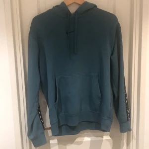 Blå hoodie från Nike - Säljer en snygg blå hoodie från Nike. Den har en stor ficka fram och en luva med dragsko. På ärmarna finns det coola detaljer och på ryggen står det 'NIKE' i svart. Perfekt för chill dagar eller träning!