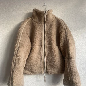 Ny Beige teddyjacka med dragkedja storlek M från H&M - Ny och oanvänd med etiketten kvar.  Tjock och varm teddyjacka i mjuk pile med kantband i ytbehandlad kvalitet. Jackan har lös passform, ståkrage, tvåvägsdragkedja fram och kängurufickor. Fodrad och lätt vadderad.  Storlek: M (något oversized) Färg: Beige, Årets trendfärg för 2025 är Mocha mousse