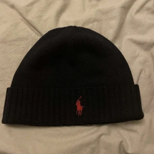 Svart mössa från Ralph Lauren - Snygg svart mössa från Ralph Lauren med röd broderad logga framtill. Den är ribbad och perfekt för kyliga dagar. En stilren accessoar som passar till det mesta.