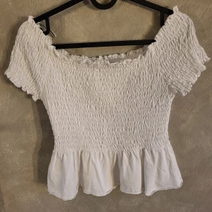 Vit offshoulder topp från Gina Tricot - Säljer en söt vit offshoulder topp från Gina Tricot. Toppen har en smockad design med volangdetaljer vid ärmslut och nederkant. Perfekt för sommarens varma dagar!