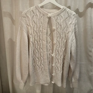 Vit stickad kofta med knappar - Säljer en egen stickad vit kofta med vackert mönster och dekorativa knappar framtill. Koftan har långa ärmar och ribbstickade kanter som ger en klassisk look. Perfekt för kyliga dagar. Den passar storlek S/M och tvättas i 30 grader, ska ej torktumlas.
