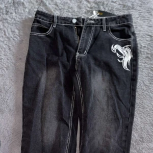 Svarta jeans med broderi - Snygga svarta jeans med vit broderad detalj på framsidan. De har en klassisk femficksdesign och kontrasterande sömmar som ger en cool look. Perfekta för en avslappnad stil.